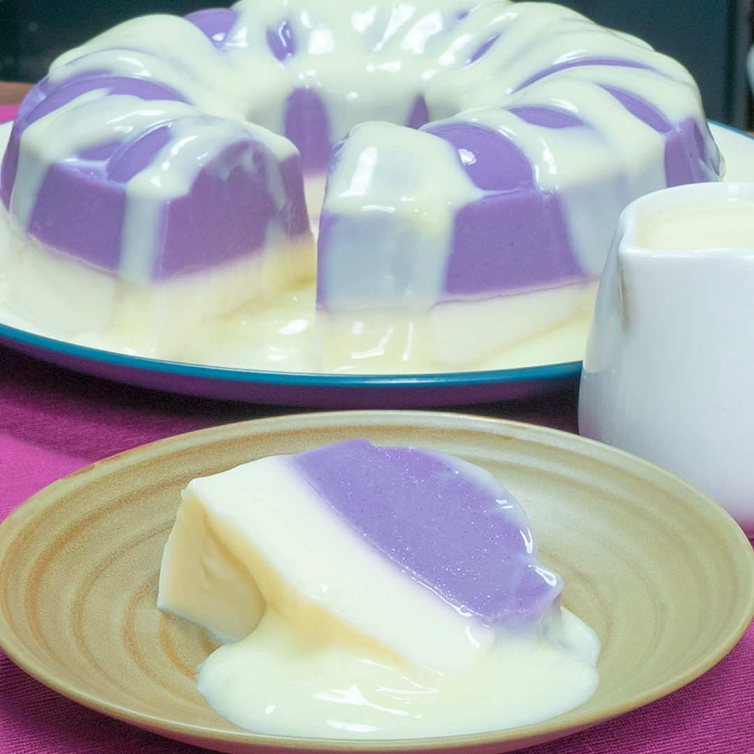 A ring-shaped purple and white layered gelatin or pudding dessert on a glass platter, with a single slice served on a tan plate alongside a small pitcher of cream.