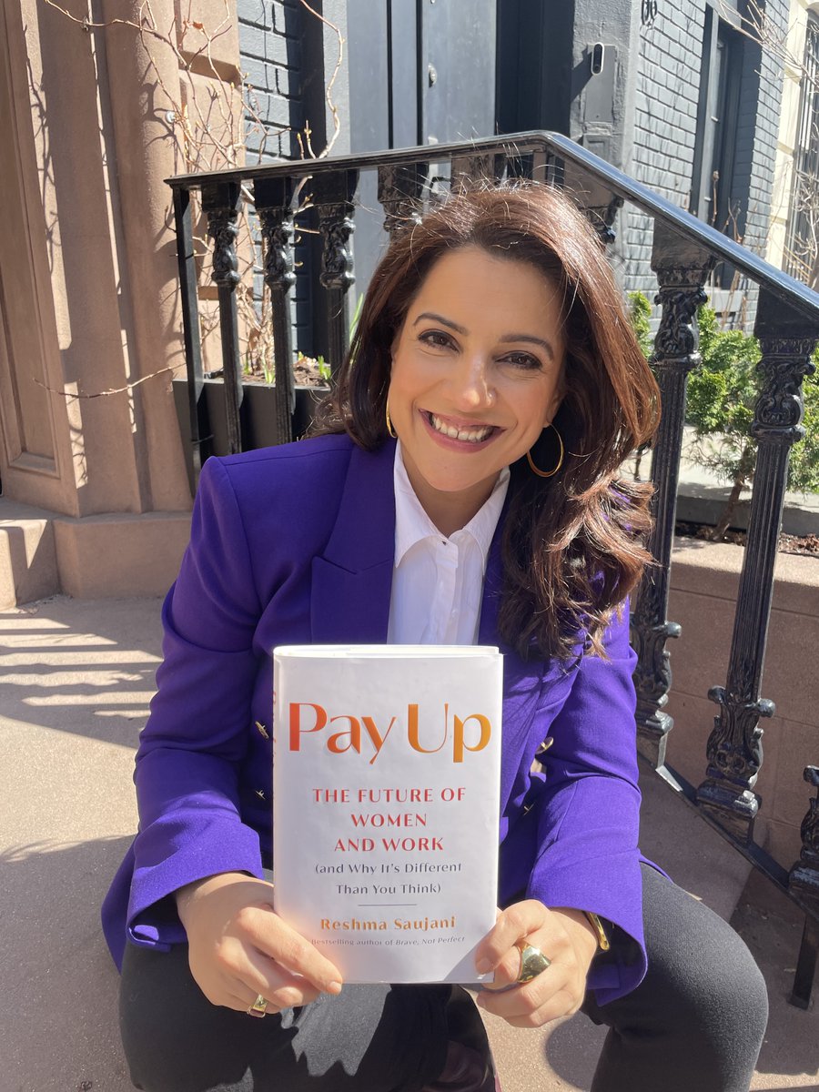 Reshma Saujani posing for a photo on a stoop holding her new book, Pay Up: The Future of Women and Work (and why it's different than you think). She is wearing a purple blazer, a white top, and black pants.