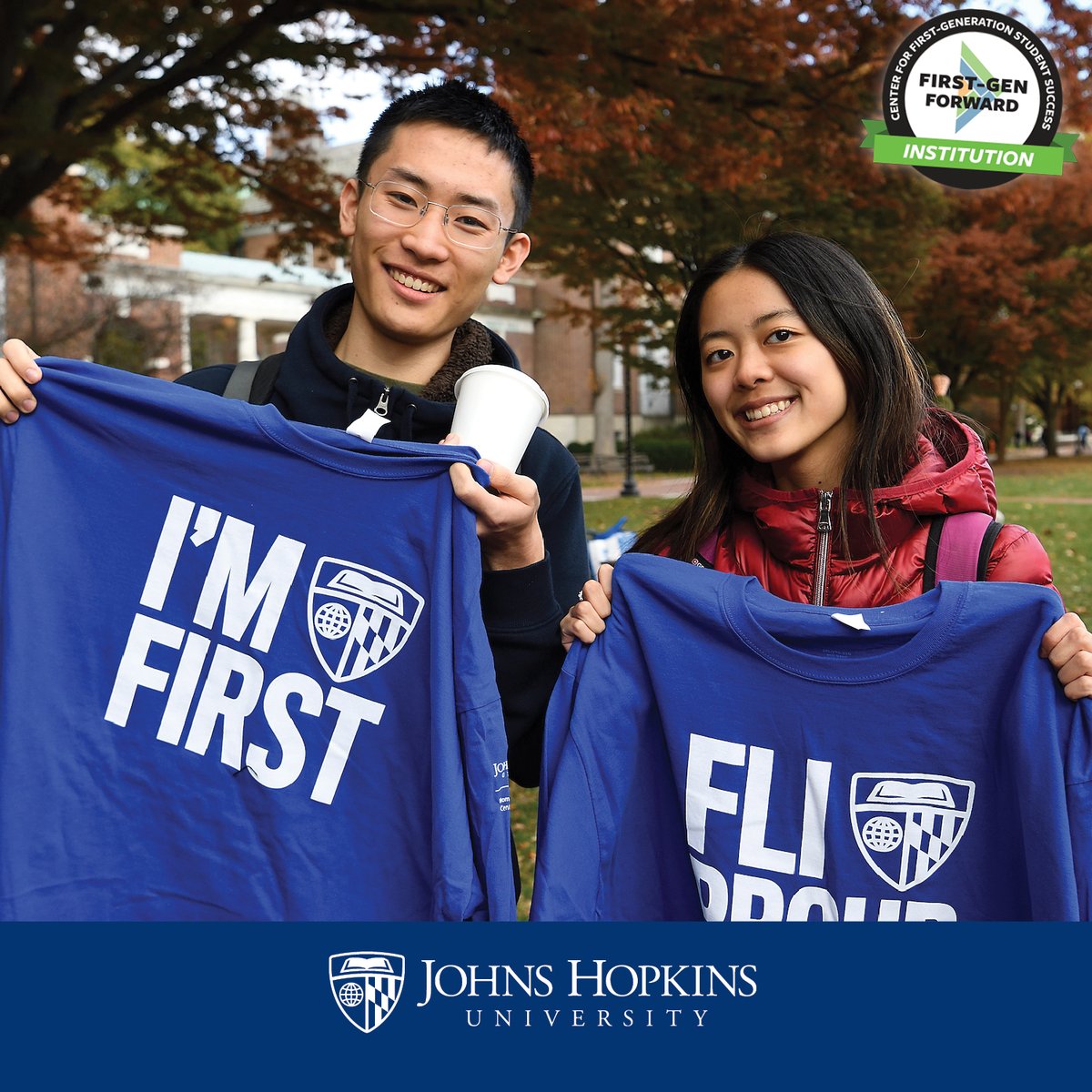 Two students stand side-by-side outdoors on the Johns Hopkins University Homewood campus, dressed for cold weather. They each smile and hold up blue long-sleeve t-shirts, one of which reads I'm First and the other that reads F L I Proud. Both shirts feature the J H U shield. In the corner is a badge logo displaying the recognition of First-Gen Forward Institution.