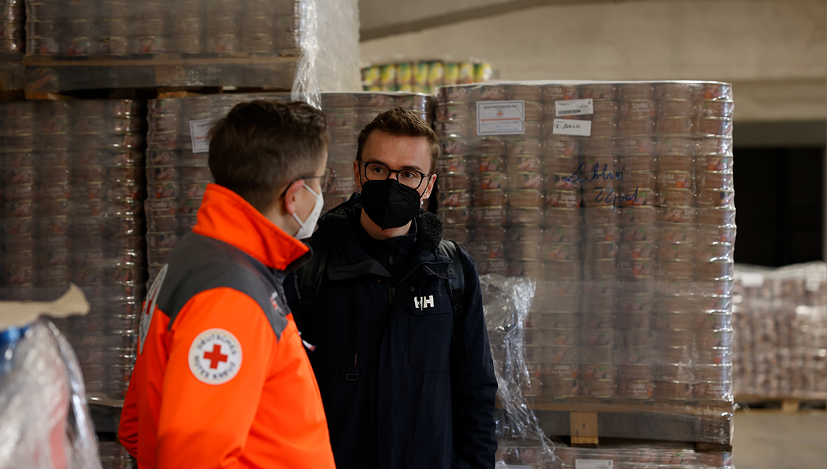 Der Grüne Bundestagsabgeordnete Julian Pahlke ist im Gespräch mit einem Mitarbeiter des Roten Kreuz zu sehen. Julian Pahlke steht frontal zur Kamera. Er trägt eine schwarze Jacke und einen schwarzen Mund-Nasen-Schutz. Der Mitarbeiter des Roten Kreuz trägt eine Uniform und einen weißen Mund-Nasen-Schutz. Sein Gesicht ist von der Kamera abgewandt und schaut Julian Pahlke an. Im Hintergrund sind mehrere Paletten mit Plastik umwickelten Konservendosen zu sehen, auf denen mit Edding Lublin geschrieben wurde.