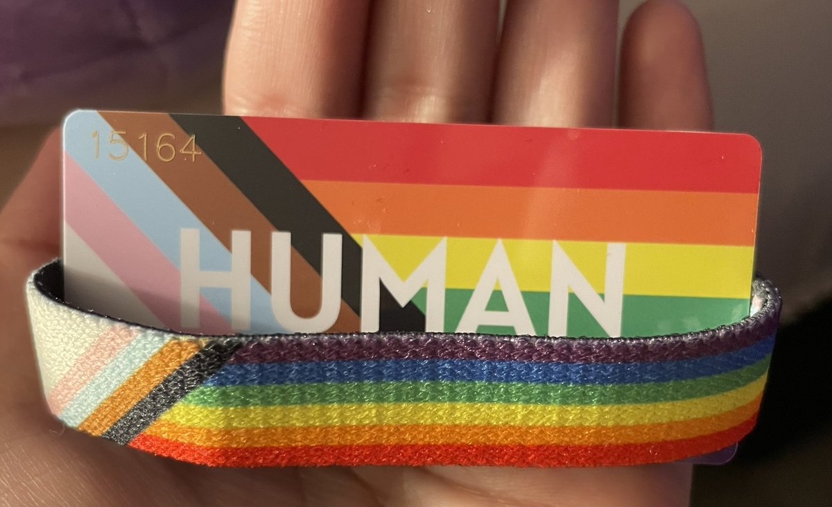 Photo of the wirst band in the palm of my hand. In the middle of the wrist band I put the little card which was sent along with it. It is striped and coloured like the progress pride flag as well, with the word “human” written in big white letters in its centre.