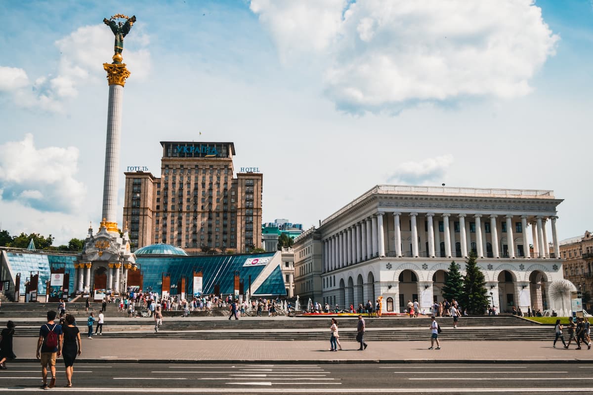 fotografia da Praça da Independência, em Kiev, capital da Ucrânia, antes da guerra. Na imagem, o dia está ensolarado e, em primeiro plano, um casal está atravessando a rua. Ao fundo, algumas pessoas estão caminhando na calçada em frente a construções históricas.