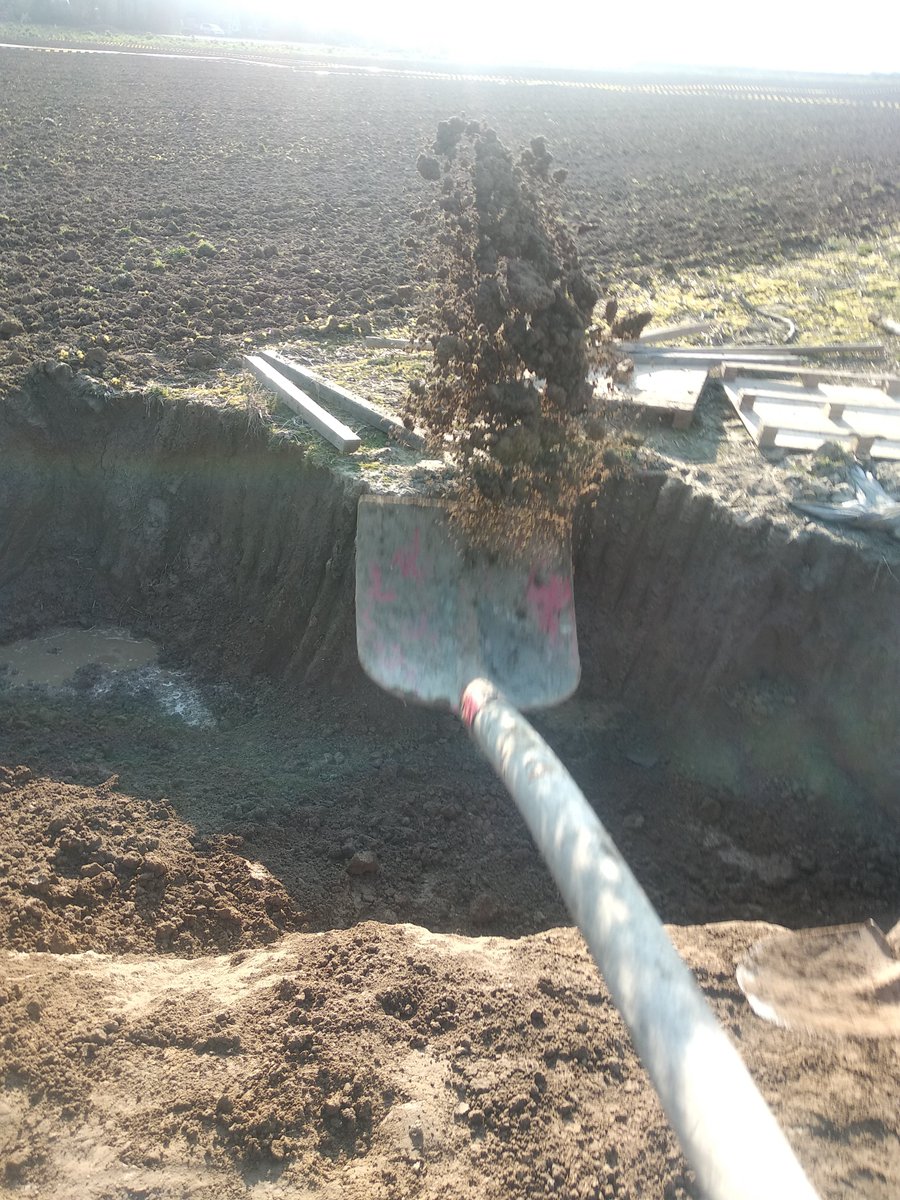 Auf dem Bild ist ein Loch am Rande eines kleinen Ackers erkennbar. Im Hintergrund ein paar Baumaterialien und am Horizont die Silhouette des Dorfes Lützerath. Blickfänger im Vordergrund ist eine eine von vorne fotografierte Schaufel mit welcher Erde in das Loch gegraben wird.