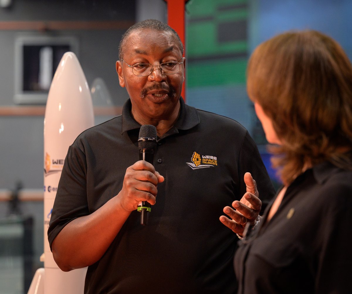 Webb Telescope Program Director Greg Robinson holds a microphone while speaking to a woman on the right. Behind him is a model of the Webb Telescope's rocket, an Ariane 5 launch vehicle.