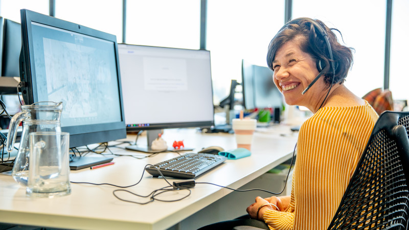 Woman wearing a headset smiling while seated at a desk with two monitors.