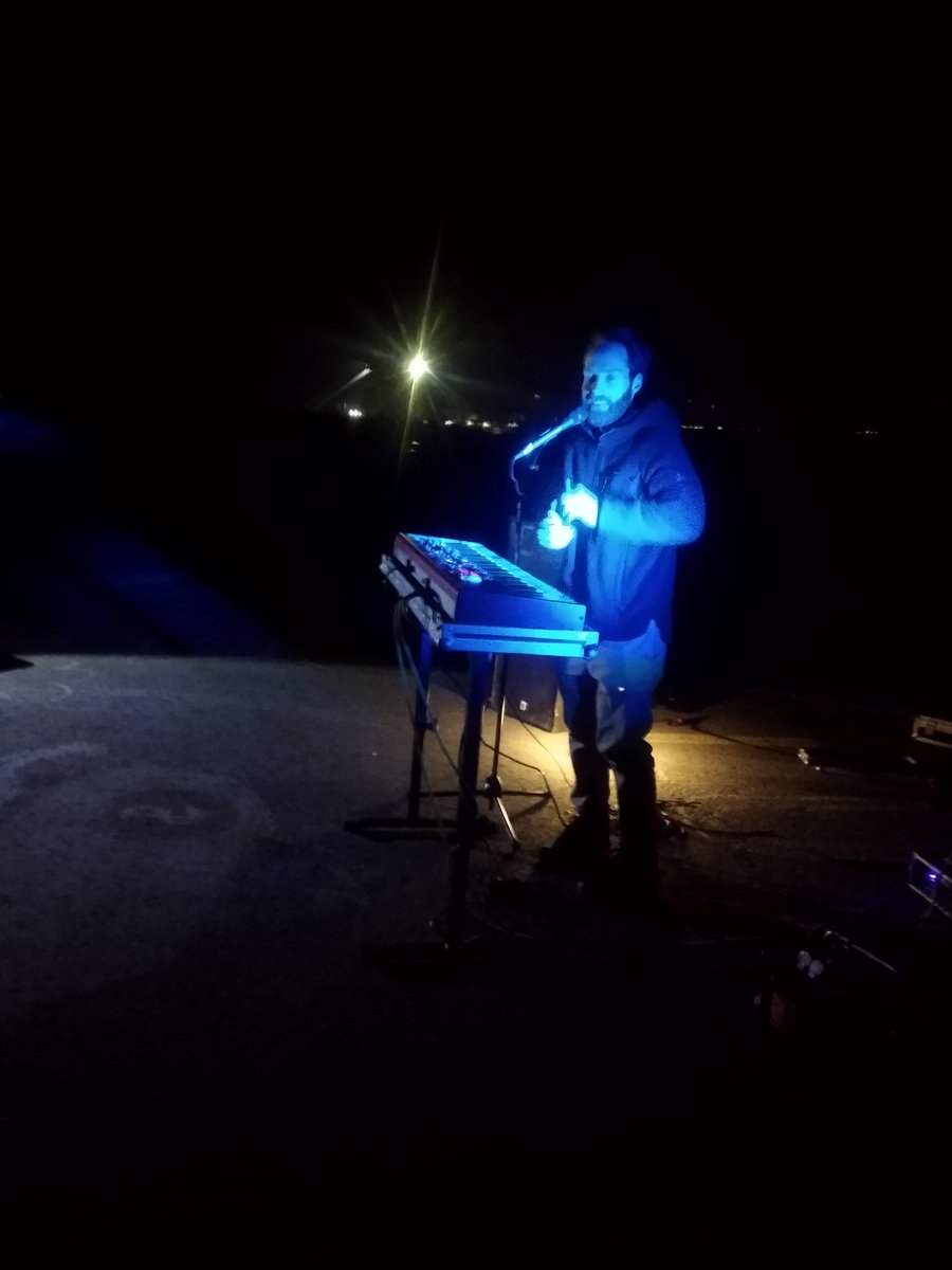 Enno Bunger im blauen Licht der Leuchtschau getaucht bei seinem Konzert in Lützerath an der Mahnwache vor der Grube. Die Erkennbaren Lichter im Hintergrund stammen von den Maschinen in der Kohlegrube. Der mit Mikrophon am Mischpult stehende Bunger blickt auf in Richtung Kamara.