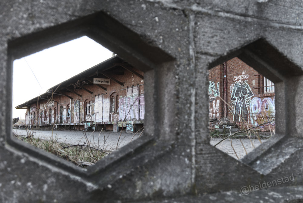 Doorkijk naar een oud goederenstation met graffiti en schildering van en naakt vrouwenlichaam. Boven een deur een bord met 'recycling'