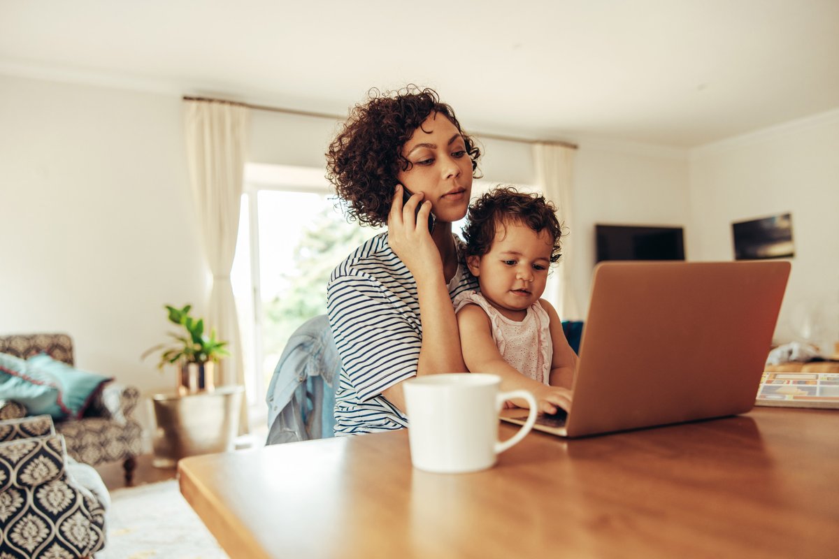 Woman sitting in front of laptop while holding a small child and on the phone at the same time. 