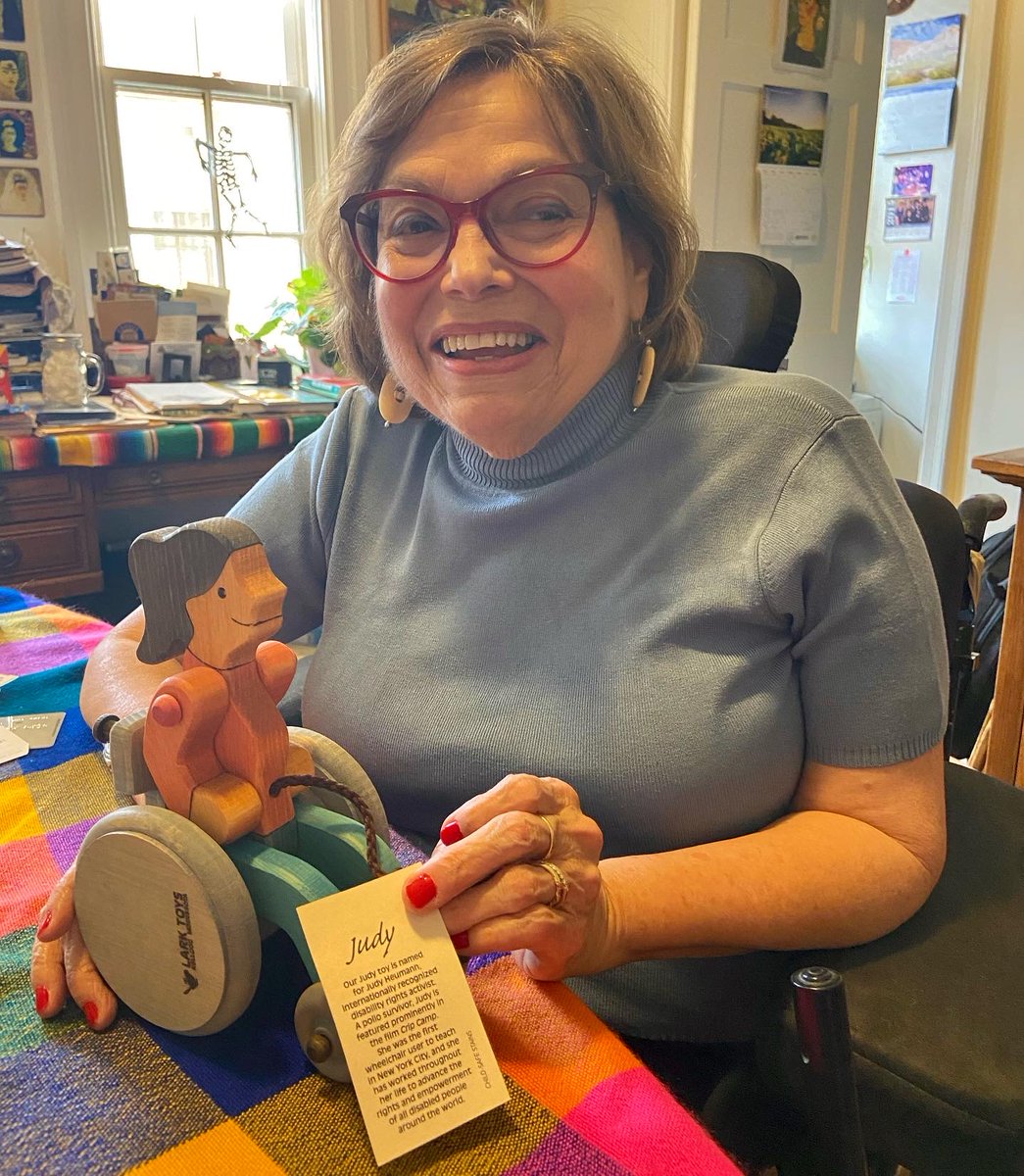 A photo of Judy Heumann smiling with a wooden toy. Judy is a white woman in her 70s who uses a wheelchair. She has short brown highlighted hair and is wearing red glasses, cream oval earrings, and a blue sweater. The toy is a girl who uses a wheelchair painted with brown hair, a pink shirt, blue pants, and a grey wheelchair. There is a brown string to pull the toy. There is a tag that reads “Judy. Our Judy toy is named for Judy Heumann, internationally recognized disability rights activist. A polio survivor, Judy is featured prominently in the film Crip Camp. She was the first wheelchair user to teach in New York City, and she has worked throughout her life to advance the rights and empowerment of all disabled people around the world.”