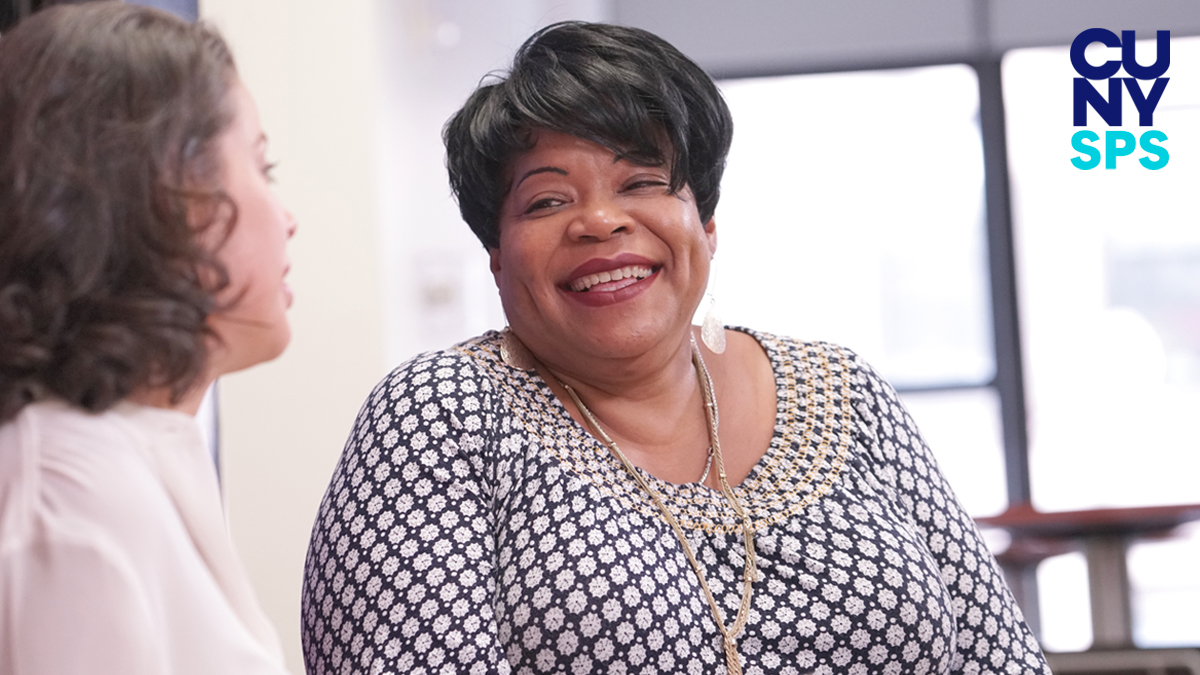 Closeup of BS in Business alum Earline Torres smiling, seated, facing fellow student and conversing, indoor setting