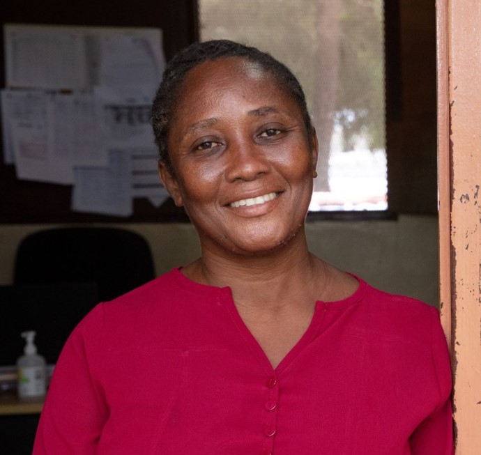 A Haitian woman wearing a magenta shirt stands in the doorway of an office, smiling.