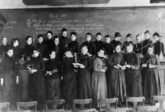 Photo of a class of students in dresses holding scientific instruments in front of a blackboard. Courtesy of MIT Museum.