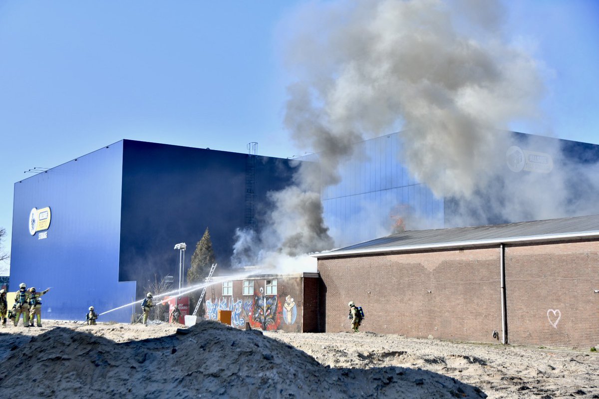 UppelMedia's tweet photo. Een gebouwtje tussen twee grote gebouwen staat in brand. Uit de naastgelegen loods komt inmiddels ook rook. Er waren meerdere vlammen zichtbaar. #Utrecht #Atoomweg https://t.co/HrHp4MIftJ