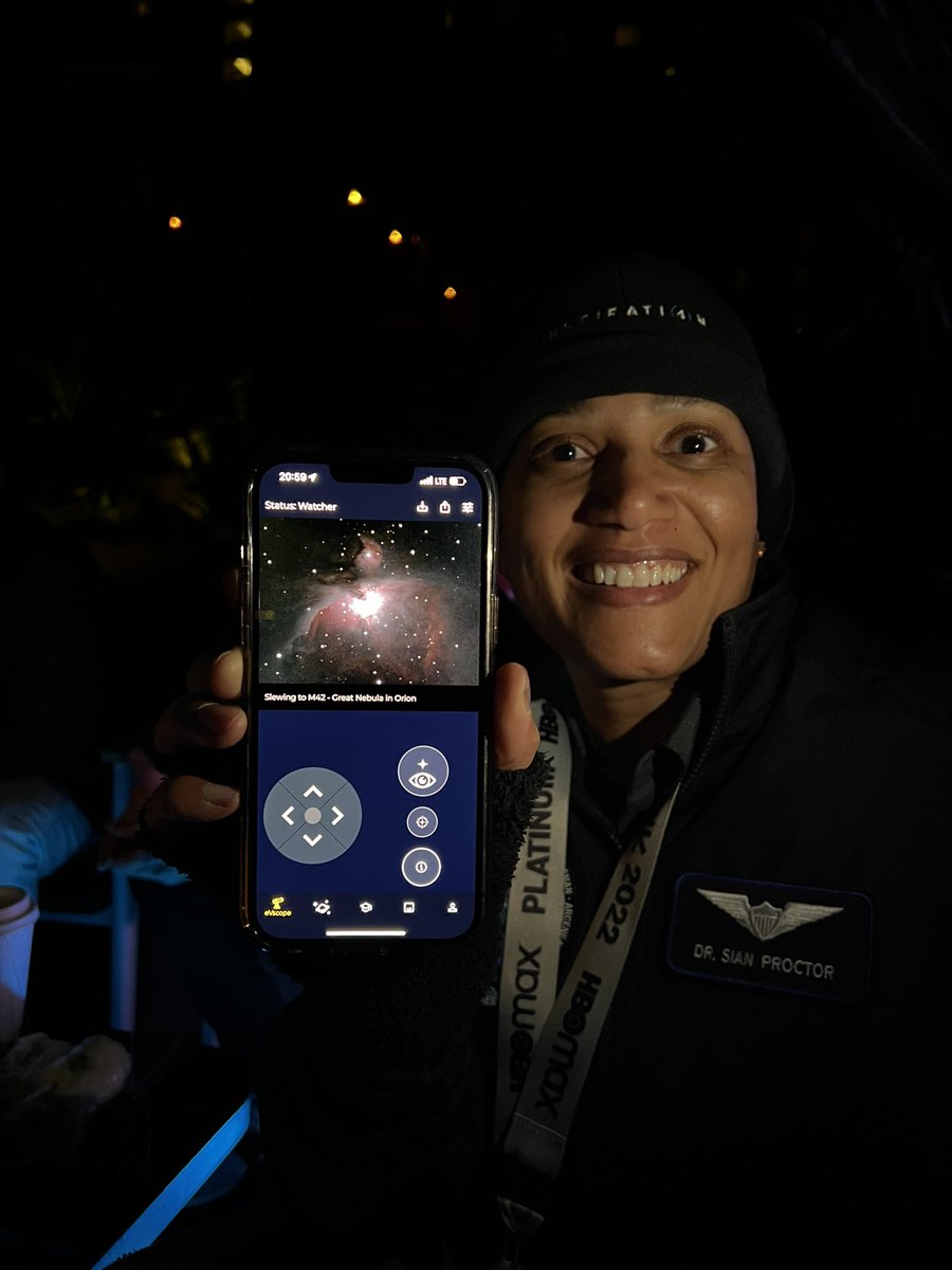 Dr. Sian Proctor holding a cell phone showing the Orion Nebula in the Unistellar app, a live view from Austin. She is smiling with a flight jacket on.