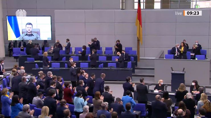 Auf dem Bild ist der Plenarsaal im Bundestag zu sehen. Auf einer Videoleinwand ist Präsident Selenskyj zugeschaltet. Die Abgeordneten haben sich von ihren Plätzen erhoben, schauen Richtung Videoleinwand und klatschen. 