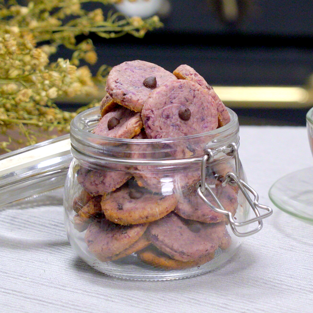 A glass jar with a clamp closure filled with purple cookies containing chocolate chips, sitting on a light-colored tablecloth with dried flowers in the background.