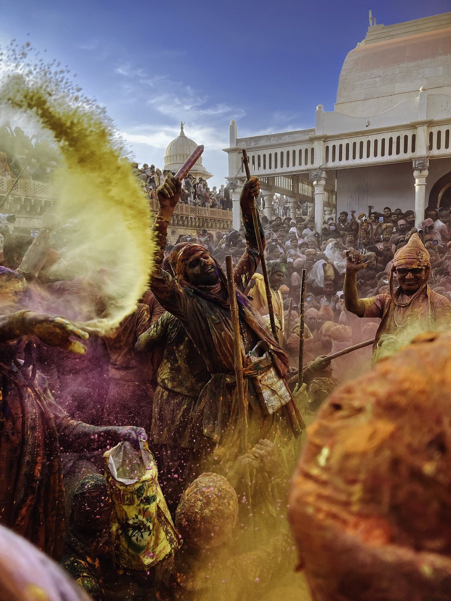 A crowded square in the town of Nandgaon, known for traditional Holi rituals, where onlookers observe a man covered in multi-colored powder with his arms raised mid-gesture.