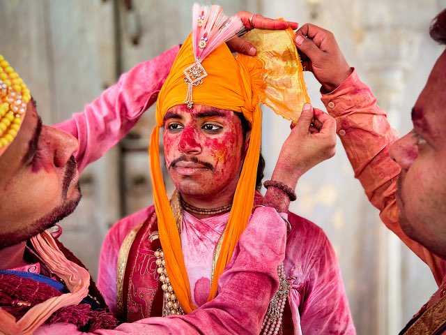 A man in the town of Barsana has two friends helping him put on a head piece made of 7 meters of yellow fabric to represent Lord Krishna.