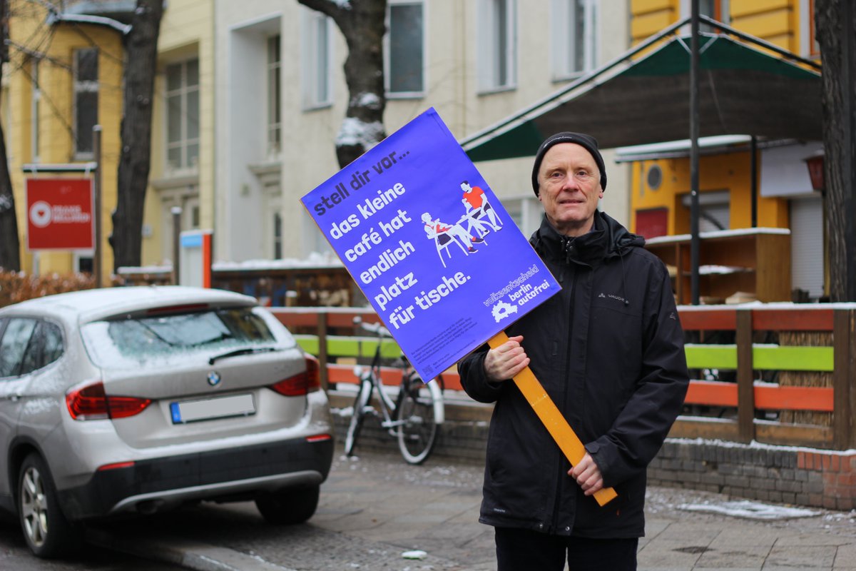 Foto von einem menschen in der Hornstraße der vor einem Restaurant steht, im Hintergrund parkt ein BMW auf dem Gehweg. Der Mensch trägt eine schwartze Vaude Jacke und eine schwarze Mütze, er selbst ist ein älterer weißer Mann und hällt ein Schild in der Hand auf dem steht: stell dir vor… das kleine café hat endlich platz für tische.” auf blauem grund mit dem Volksentscheid Berlin Autofrei Logo unten rechts
