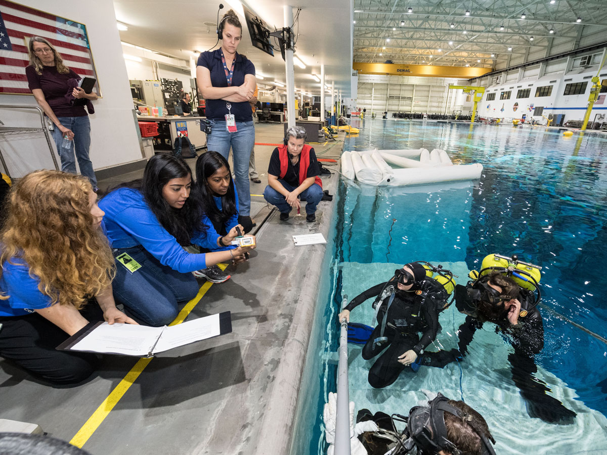 Interns, all women, standing at the edge of a training pool at Johnson Space Center with divers inside. The interns have notebooks and are taking notes. Two are of color, one is a redhead.