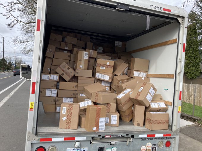 The back of a uhaul box truck is opened and piles of cardboard boxes are stacked in the back.