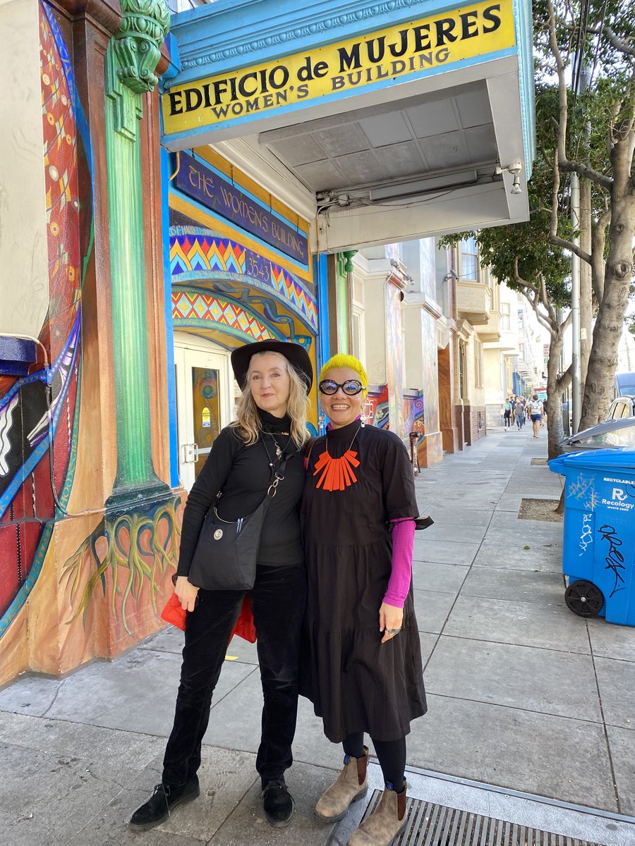 Rebecca (left) with her arm around Mona, standing on a pavement under a sign that says Edificio de Mujeres. Rebecca is wearing black trousers, black top, black jacket and black hat. Mona is wearing a black dress with magenta sleeves visible underneath.