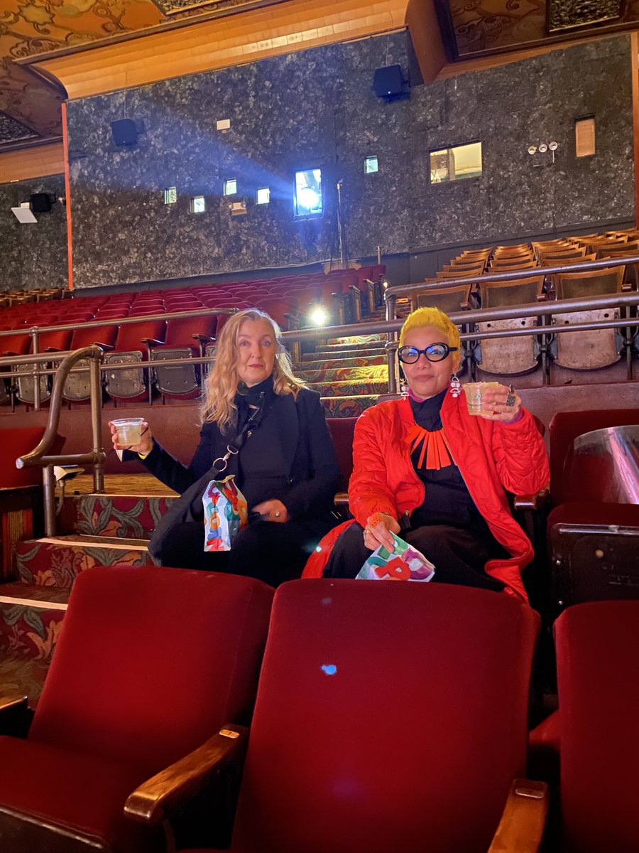 Rebecca (left) sitting next to Mona on red seats surrounded by other empty red seats in a movie theatre. They are holding up plastic cups full of a clear liquid. Rebecca is wearing a black top and jacket and Mona is wearing an orange coat over a black dress.
