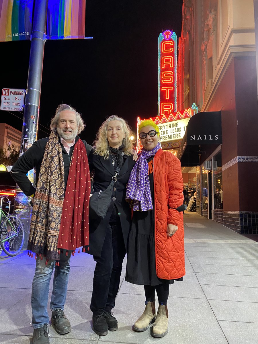Robert, Rebecca, and Mona standing on a pavement and visible behind them is a marquee that says Castro and the name of the film Everything Everywhere All At Once. Robert is wearing blue jeans and a black top and a red and brown scarf. Rebecca is wearing black trousers and a black jacket. Mona is wearing an orange coat over a black dress and a purple and white scarf.