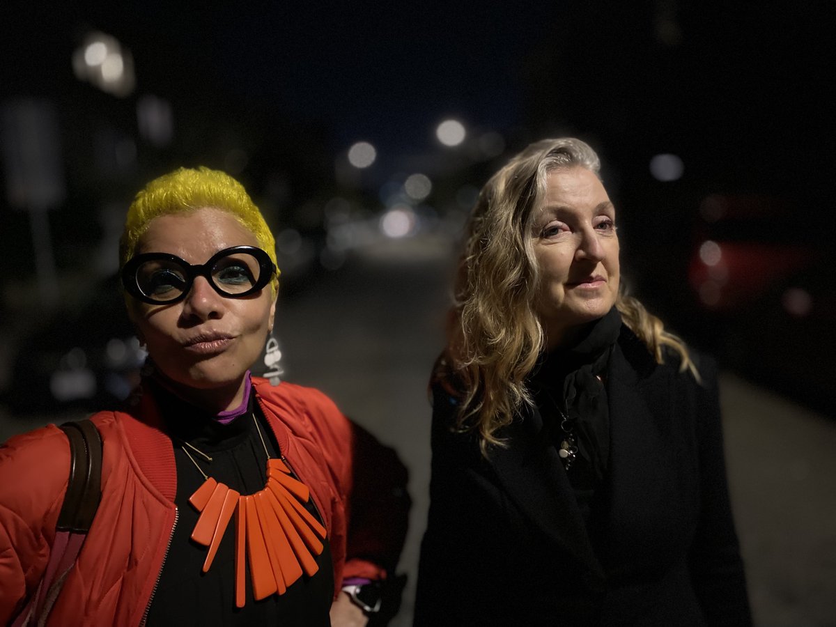 Mona (left) and Rebecca from the shoulders up. Mona is wearing an orange coat over a black dress and an orange necklace.   Rebecca is wearing black coat over a black top.