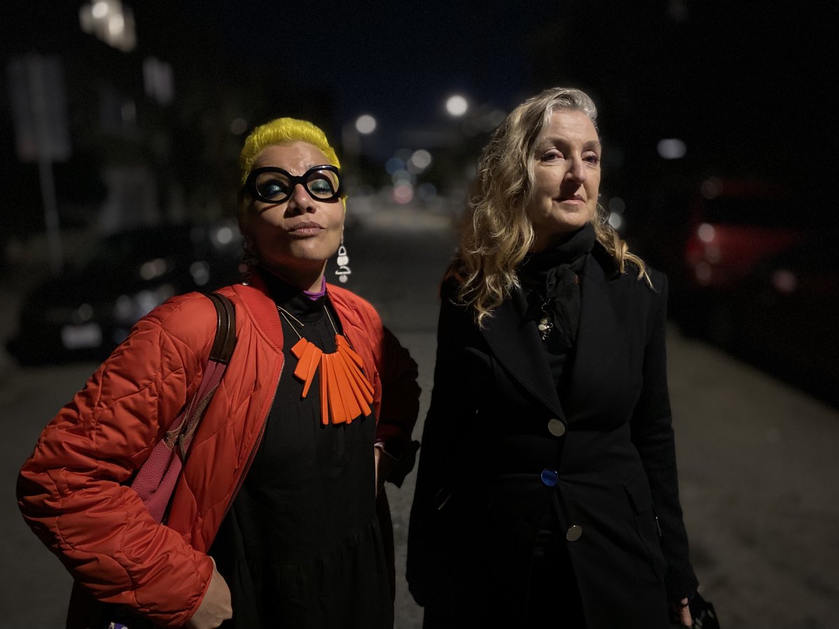  Mona (left) and Rebecca from the waist up.. Mona is wearing an orange coat over a black dress and orange necklace. and her right arm is on her hip. Rebecca is wearing a black coat over a black top.