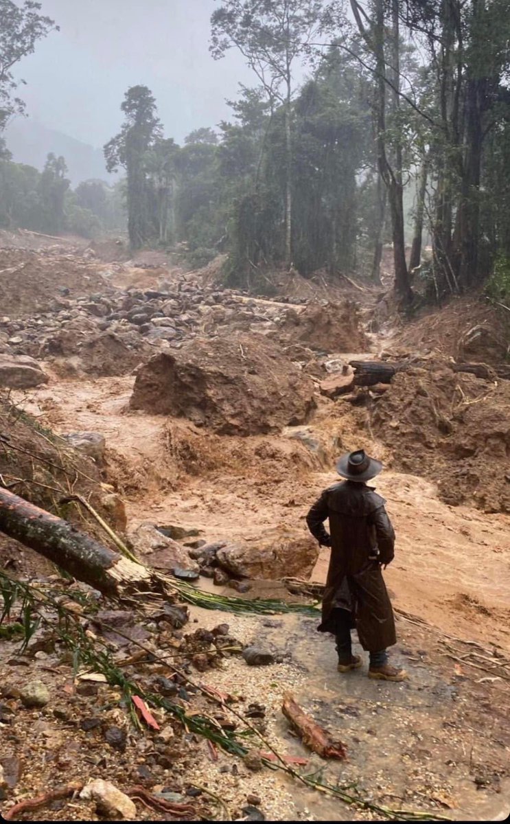 Man in hat, long rain coat and heavy boots watches heavy rain causing muddy river to destroy part of forest.