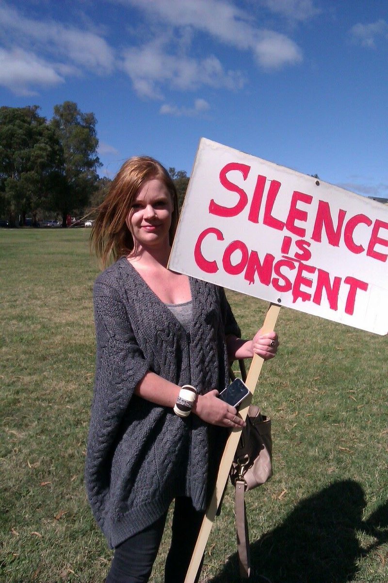 Young woman holding a wooden stake bearing a sign saying Silence is Consent, letters in red on a white background