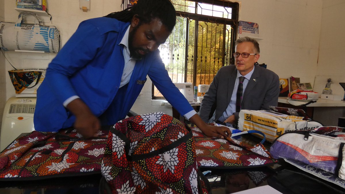 Innocent demonstrates to Balozi @DConcar one of the innovative creations at Soma; a suit storage bag made of recycled fish food sacks. @UKinTanzania supported Soma in equipping new premises where thousands of bags will be made to help the community & clean up the environment.