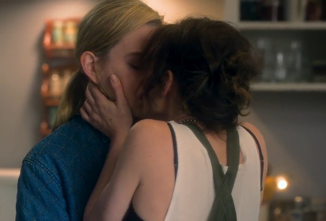 A scene showing two women kissing intimately, one wearing a blue denim top and the other in a white tank top with olive green overall straps, in what appears to be a warmly lit kitchen setting.