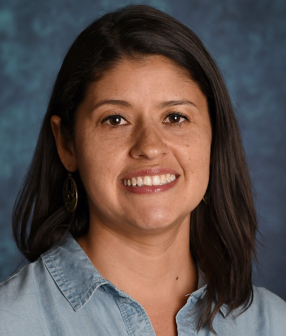 Portrait of Dr. Ivette Guzmán. She has long dark hair, brown eyes, and a big smile. She is wearing a light blue shirt with a collar in front of a blue background.
