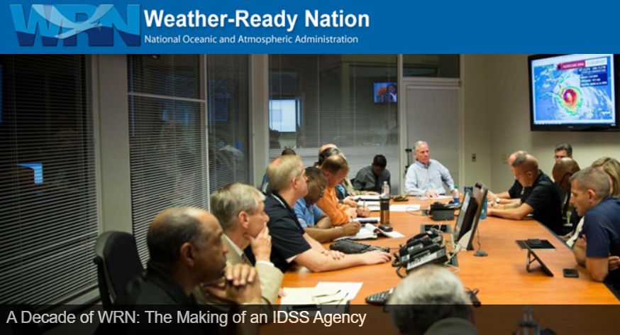 Photo of an emergency operations briefing with officials around a conference table and hurricane information on a TV screen.