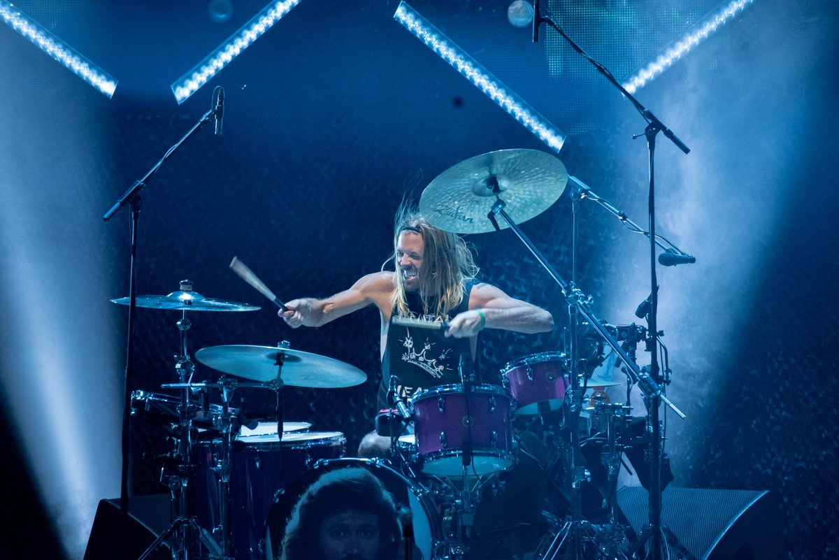 A photo of Taylor Hawkins on drums. His hair is down and he has a passionate grimace on his face. Both arms are extended as he plays the drums.