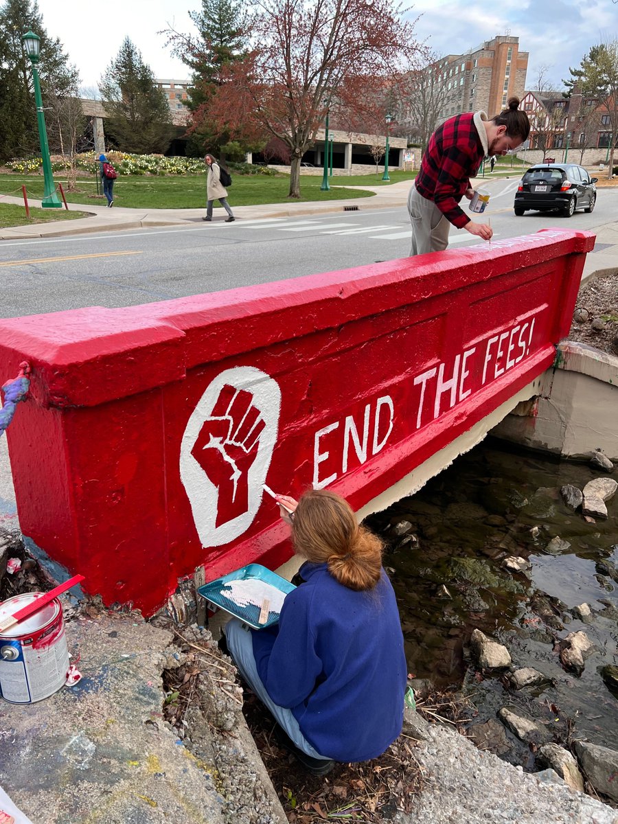 Two people paint a red bridge with the solidarity fist and the words "END THE FEES"