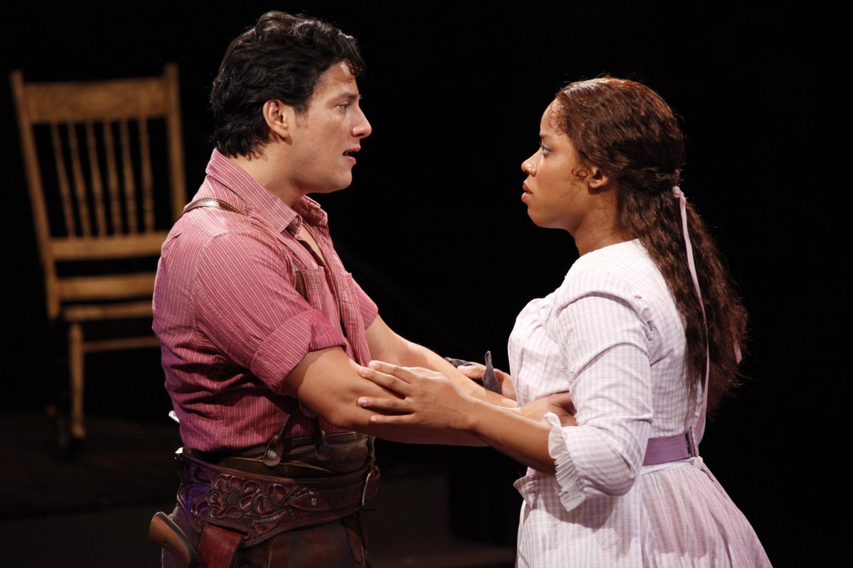 Nicholas Rodriguez as Curly and Eleasha Gamble as Laurey in the Arena Stage at the Mead Center for American Theater production of Rodgers and Hammerstein’s Oklahoma! Photo by Carol Rosegg. A man in a pink shirt and a leather gun belt holds a woman in a pink blouse and skirt. They look into each other's eyes.