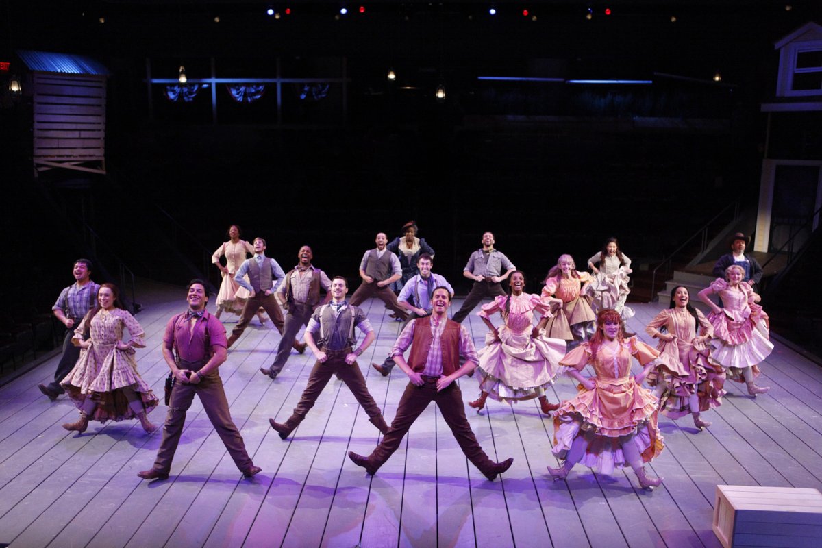 The company of the Arena Stage at the Mead Center for American Theater production of Rodgers and Hammerstein’s Oklahoma! Photo by Carol Rosegg. A group of actors in turn-of-the-20th-century cowboy garb and colorful dresses dance facing the audience. They are all posed with their legs wide, balancing on their heels.
