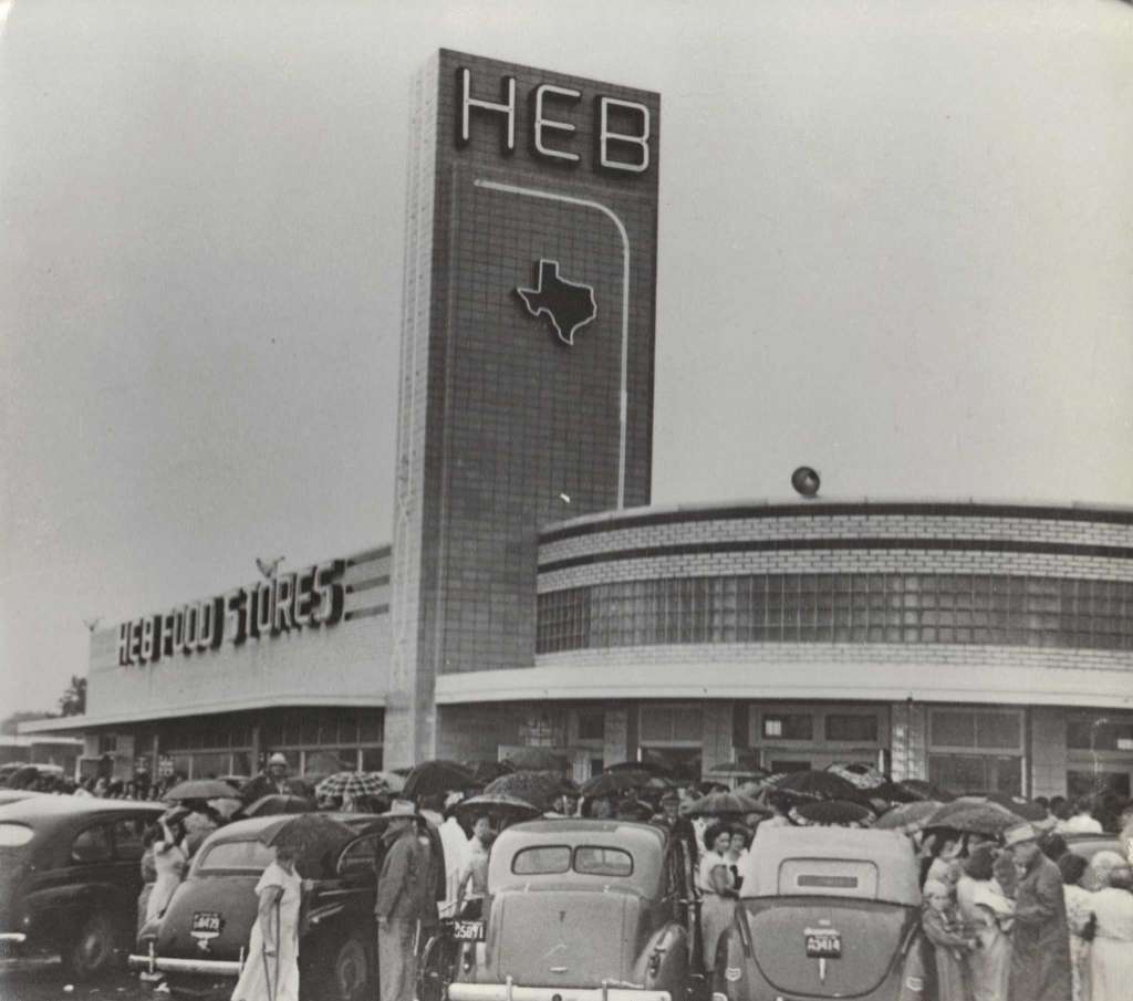 Vintage black-and-white photo of an H-E-B grocery store with a tall sign and a curved glass-block façade, its parking lot full of 1940s-era cars and people holding umbrellas