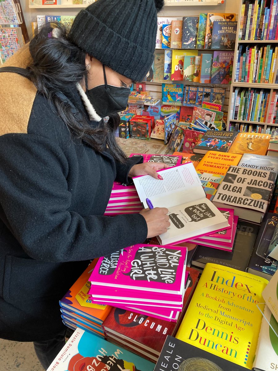 An action shot of Julissa signing copies atop a display table, with lots more books in the background.