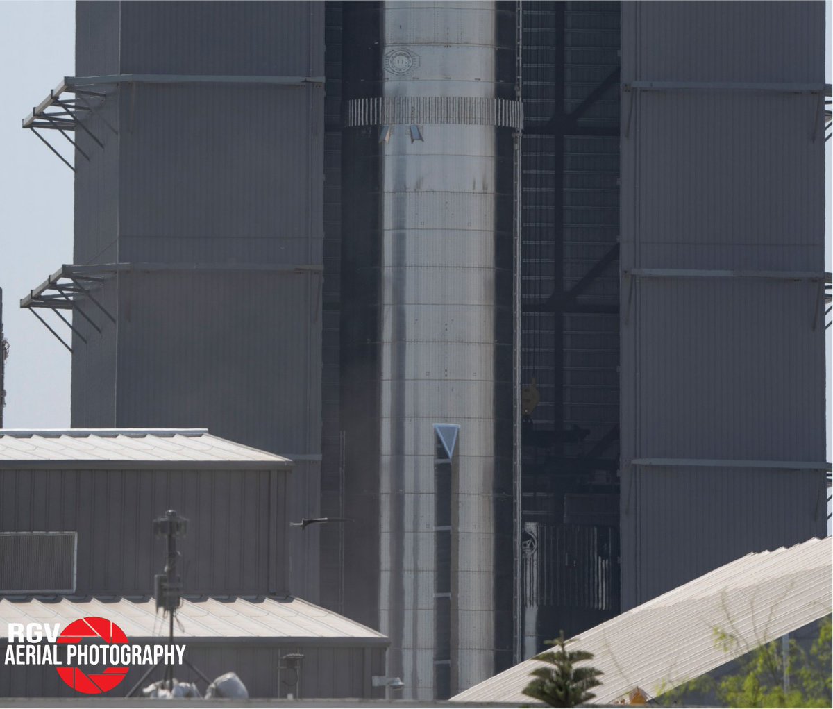 CSI_Starbase's tweet photo. (1/2) Hey #StarshipAddicts, it appears #Booster7 has a company in the #Highbay. Earlier today @RGVaerialphotos caught a picture of #Booster8 forward section tucked almost out of sight! If you look closely you can see the #BridgeCrane load spreader preparing to attach to it. https://t.co/Ape5yXbqMl