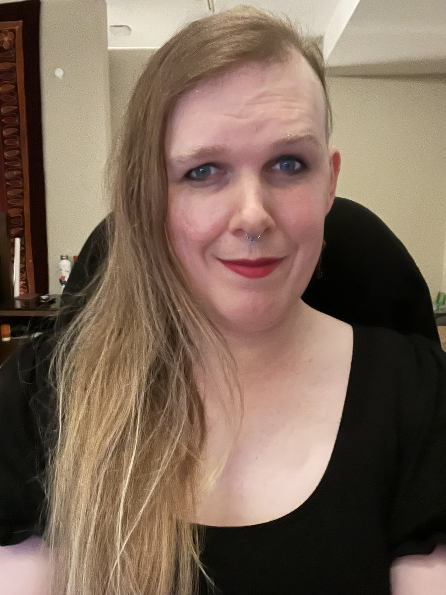 White trans femme with dirty blonde hair and blue eyes wearing a black scoop neck dress. She’s wearing some dark mascara, red lipstick, and has a septum piercing.