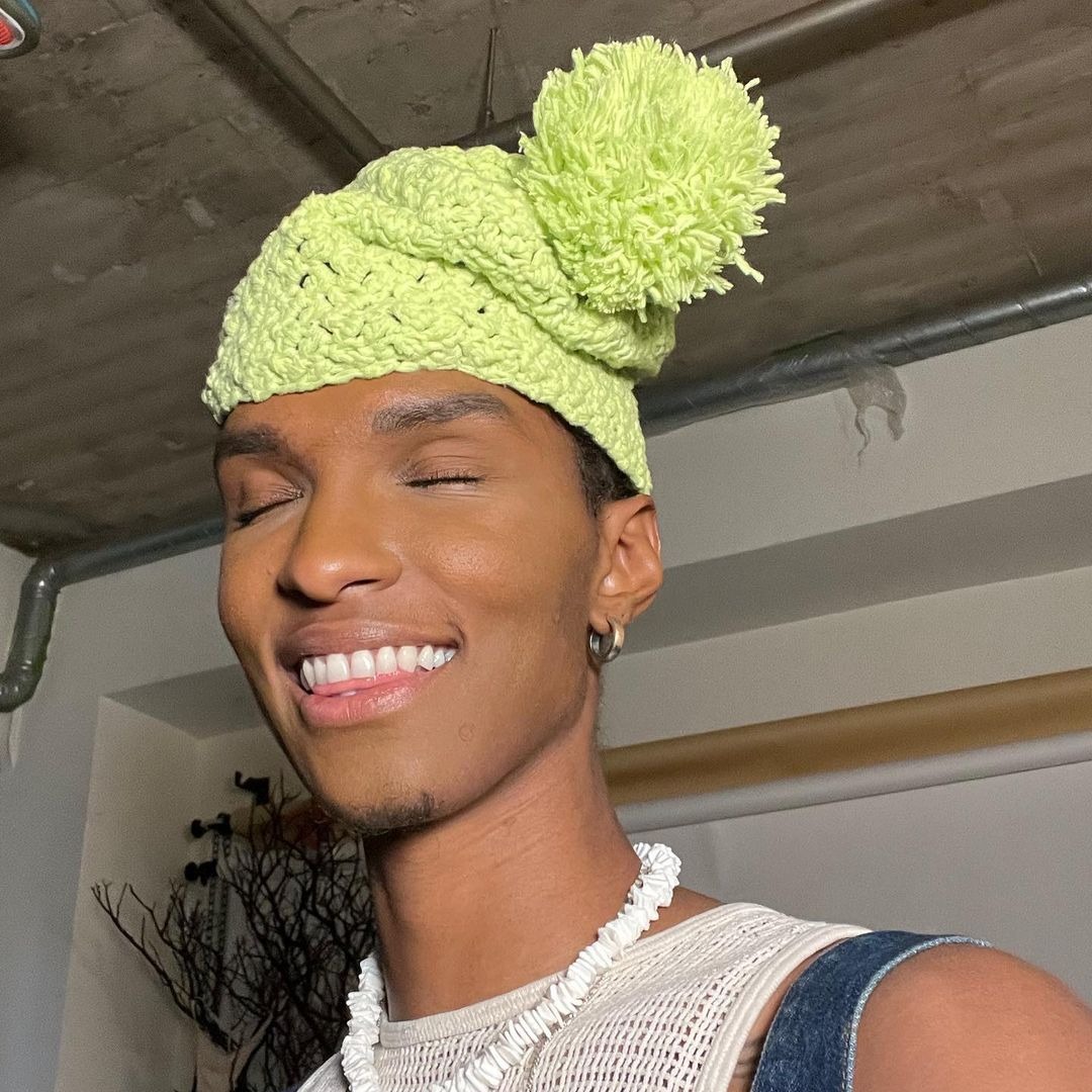 A picture of creator ARROWS smiling, eyes closed to camera, wearing a bright green knit hat, a shell necklace and chains, over a white-knit tank top and blue overalls.