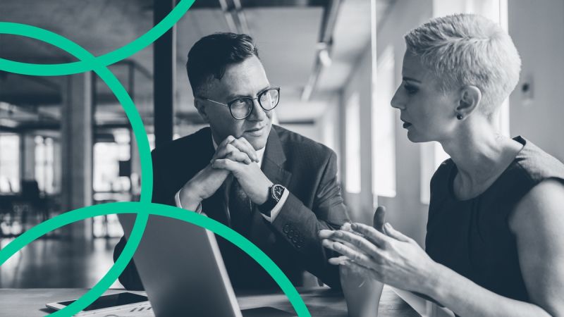 Male and female sitting at a table, facing each other, with a laptop open in front of them. Male has clasped hands and female has her hands raised off the table in a gesture to explain something.