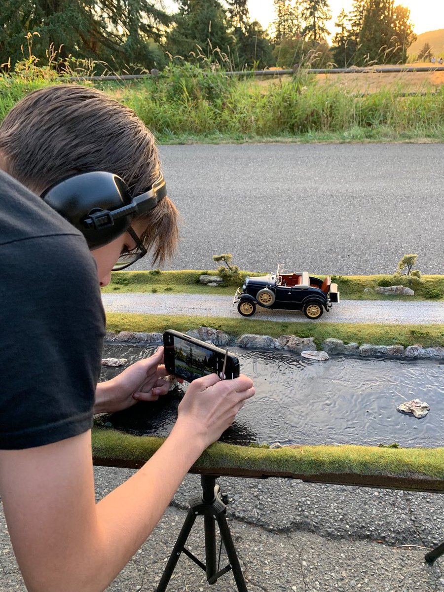 An over-the-shoulder photo of Anthony taking a photo using his iPhone on a tripod. Anthony wears over-the-ear headphones and aims his iPhone camera at a small, black, old-time model car parked on a miniature roadway situated behind a small flowing river. Beyond the roadway is a life-sized road, with the sunset peeking over a meadow in the distance.