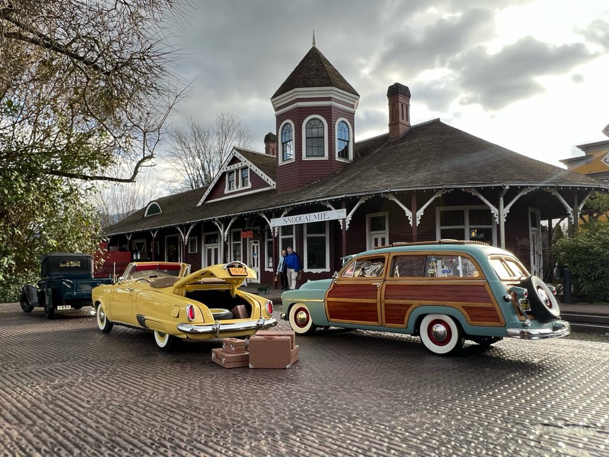 Three miniature old-time model cars parked in a cobblestone drive in front of a business complex. The cars appear to be life-sized and two people are seen in the distance walking toward the vehicles. One yellow vehicle has its trunk open with luggage spilling out of the back.