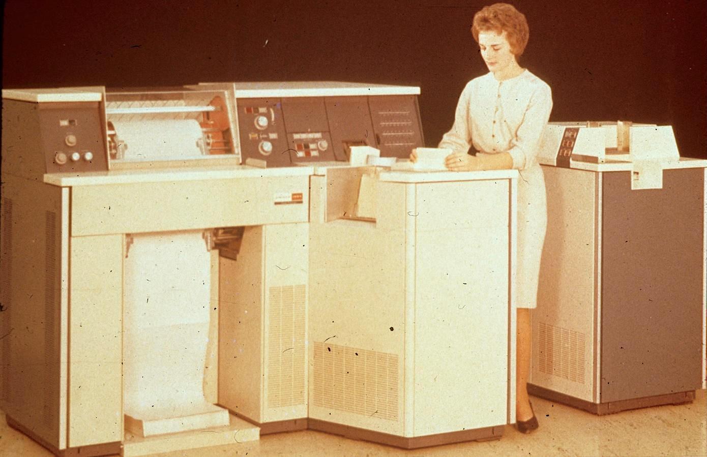 Vintage photo of the Univac 1004 with a woman using it. The main part of the system is a line printer, with a couple of dishwasher-sized cabinets attached.