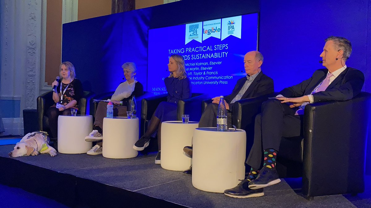 Five panellists on a stage, in the order of three women and two men. The two women on the left are holding a microphone.